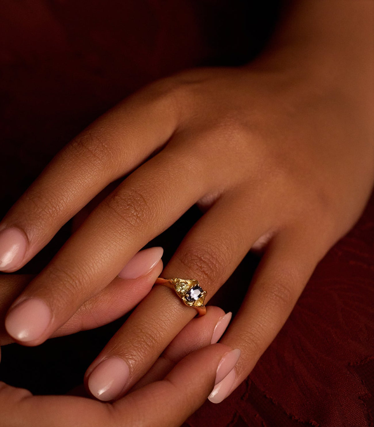 A close-up of a hand wearing a gold ring with a small sapp gemstone, resting over another hand with pale pink manicured nails against a dark red textured background.